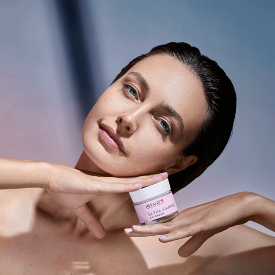 Žena drží pod bradou nádobu s ružovým nápisom „Extra-Firming Day Cream“ a rukami si jemne rámuje tvár.